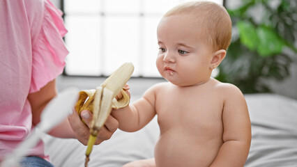Loving mother sitting on bed, sharing a healthy banana snack with her daughter, binding their...