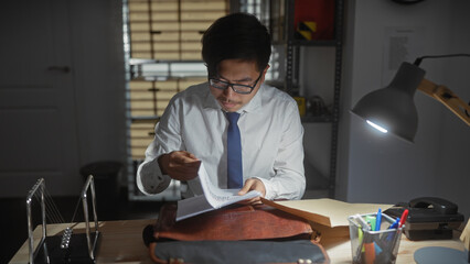A focused asian man reviews documents at his office desk, portraying an investigatorâ€™s...