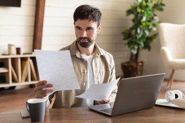 A man is seated at a table, working on a laptop and surrounded by papers. He appears focused and...