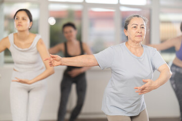 Poised senior woman, dressed in comfortable t-shirt and leggings, executing dance moves with focused precision among group of younger female ballet amateurs rehearsing in studio..