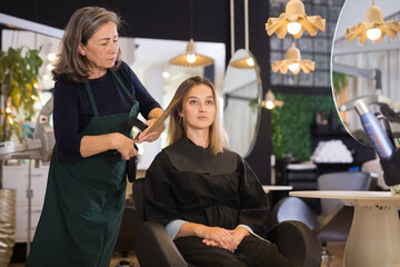 Attractive brown-haired young woman getting haircutting by professional elderly female hairdresser...