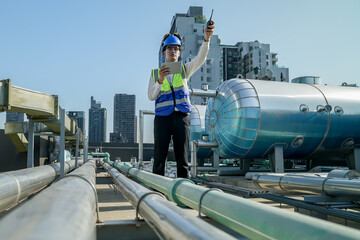 Naklejka premium A young engineer checks equipment on a rooftop amidst urban skyline, clipboard in hand, ensuring system functionality.Engineer in safety vest and helmet conducting maintenance on rooftop machinery.