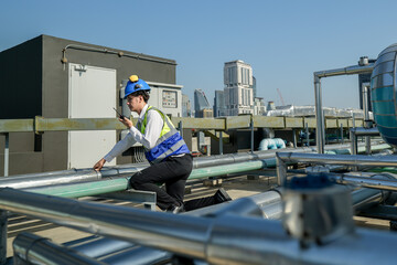 An engineer attentively checks pipework on a rooftop against a cityscape, using a tablet for data...