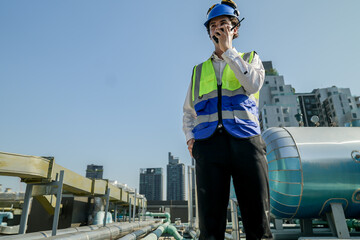 A young engineer checks equipment on a rooftop amidst urban skyline, clipboard in hand, ensuring system functionality.Engineer in safety vest and helmet conducting maintenance on rooftop machinery.