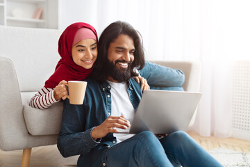 Cozy home scene of Muslim couple exploring the web on a laptop, surrounded by books and personal...
