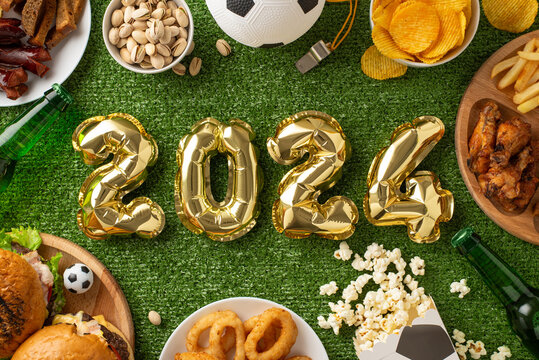 Euro 2024 soccer viewing setup. Top view shot of 2024-shaped balloons, party snacks like chips, nuts, fried chicken, onion rings, popcorn, chips, beer bottles, football, referee whistle on grass field - Powered by Adobe