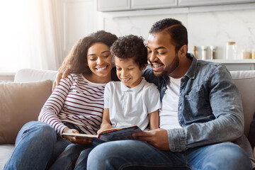 Black family consisting of parents and children are seated together on a couch, engrossed in...