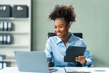 Utilizing a tablet, a young African American woman in a blue formal shirt with afro brown hair...