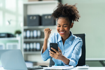 Exuberant and triumphant, a young African American woman in a blue formal shirt with afro brown hair operates a tablet, notebook, and mobile phone in a modern office setting.