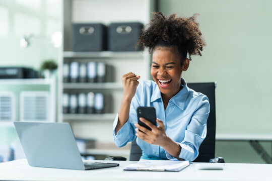 Exuberant and triumphant, a young African American woman in a blue formal shirt with afro brown hair operates a tablet, notebook, and mobile phone in a modern office setting.