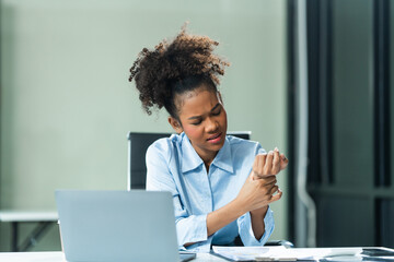 A young African American woman in a blue formal shirt with afro brown hair battles office syndrome, including wrist pain, repetitive stress, arthritis, and carpal tunnel syndrome in a modern office.