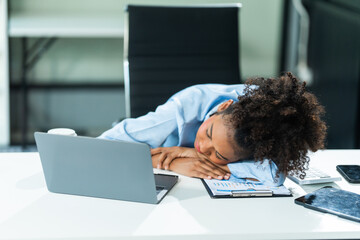 A young African American woman in a blue formal shirt with afro brown hair experiences office syndrome, feeling tired and sleepy, sometimes resorting to taking naps at her desk in a modern office.