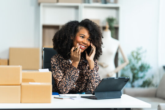 A young African American woman with afro brown hair works in a modern office, managing her online clothing store and live stream platform, sme box packages.
