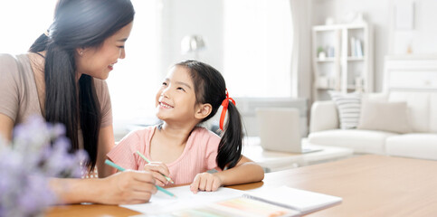 Asian mother and her small daughter sitting in cozy living room, teaching girl paint use album and colourful pencils