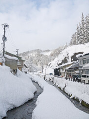 Beautiful Japanese small village winter scenery with traditional wooden houses with big snowdrifts on the roofs, river, pine forest in snow on the hill, Ginzan onsen hot spring, Yamagata, Japan