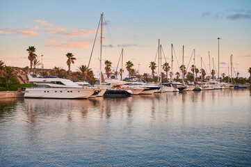 Yachts and sailboats docked in a marina harbor in the Mediterranean Sea at sunset.