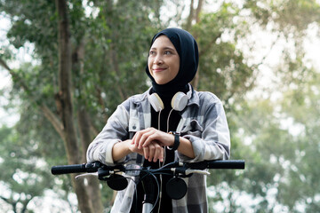 Fototapeta premium Young cheerful hijab woman, wearing casual flannel riding bicycle bike on sidewalk in the city park enjoying being outdoors, look aside or ahead. People active urban healthy lifestyle cycling concept.