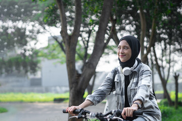 Young cheerful hijab woman, wearing casual flannel riding bicycle bike on sidewalk in the city park...