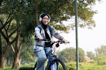 Fototapeta premium Young cheerful hijab woman, wearing casual flannel riding bicycle bike on sidewalk in the city park enjoying being outdoors, look aside or ahead. People active urban healthy lifestyle cycling concept.