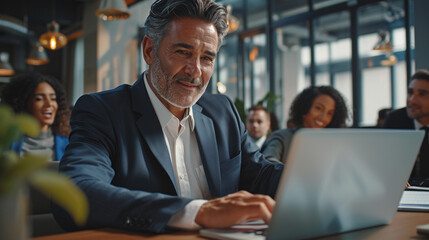 In a modern corporate setting, a successful middle-aged businessman works diligently on his laptop at his desk. He is surrounded by a diverse group of happy professionals in the multi-ethnic workplace