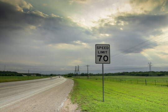 On a tranquil Texas highway, a "70" speed limit sign demands notice, flanked by roadside grass and distant power lines vanishing into the horizon. As the dark clouds of a spring storm disperse.