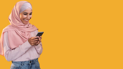 Mobile chat. Online communication. Social media. Happy smiling woman in hijab using phone texting message isolated on orange empty space background.