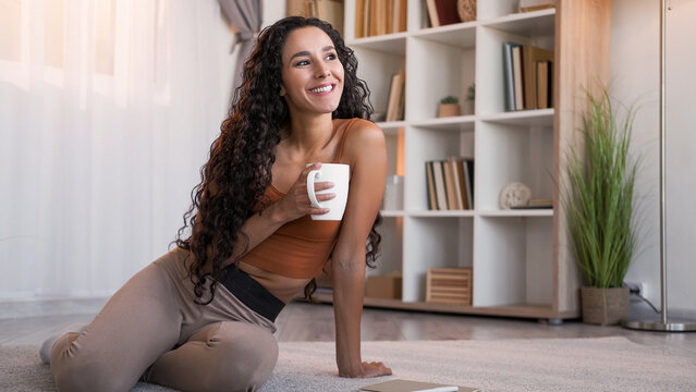 Morning coffee. Home leisure. Weekend rest. Happy relaxed smiling woman with mug daydreaming enjoying hot drink on floor at modern living room interior with free space.
