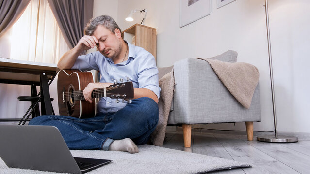 Guitar learning. Educational problem. Virtual lesson. Upset displeased man sitting on floor studying at laptop video course playing chords in home interior copy space.