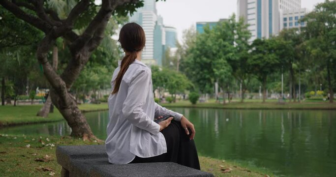 Thoughtful woman on park bench near lake skyscrapers look behind side view relaxation in close-up. Relaxation in park where city meets nature Relaxation in park tranquil contemplation.