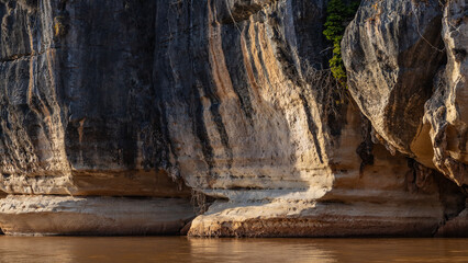 Steep coastal karst cliffs. Limestone sheer slopes rise above the red-brown river. Green plants on...