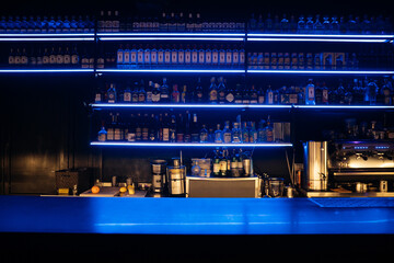 A bar with a blue counter and shelves of liquor. The bar is empty and the lights are on.