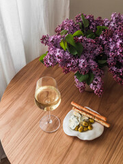 A glass of white wine, a plate with snacks, a bouquet of lilacs on a wooden table, top view