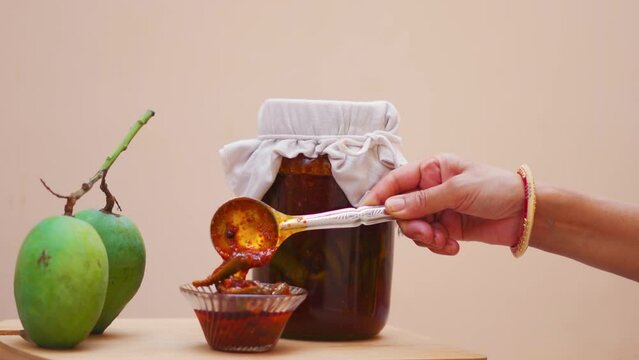 Close up shot of Homemade Mango Pickle or aam ka achar in bowl with raw mangoes and transparent jar in background. Indian homemade raw mango pickle. Indian woman hand with spoon of pickle. 