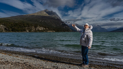 A man in a down jacket stands on the shore of a beautiful emerald lake. The hand is raised. The waves are foaming on the pebbles. Picturesque Andes mountains against a blue sky and clouds. Lago Roca. 
