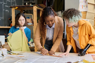 Portrait of Black young woman teaching two girls with disabilities sewing clothing in inclusive...