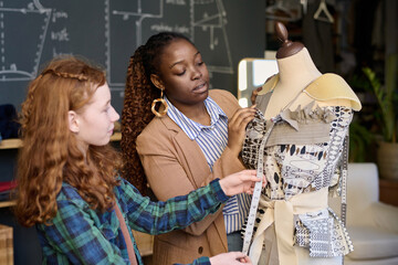 Waist up portrait of teenage girl with female teacher fitting designer clothes to mannequin in...