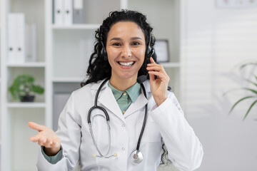 A cheerful Hispanic woman dressed as a doctor communicates via headset in a bright, modern clinic...