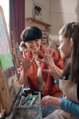 Vertical portrait of playful young woman as female teacher in art class for children showing hands...