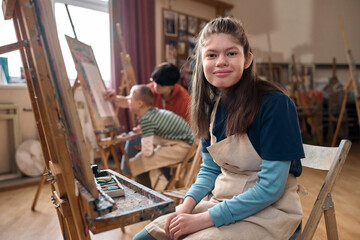 Portrait of teen girl with disability smiling at camera sitting by easel in art class copy space