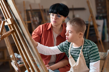 Side view portrait of young boy with Down syndrome painting on easel in art class enjoying creative...