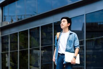 Portrait of handsome Asian student holding computer laptop and backpack. A young man standing...