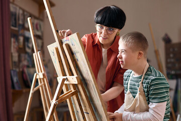 Side view portrait of smiling boy with Down syndrome sitting by easel in art class with teacher...