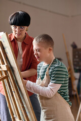 Vertical portrait of smiling boy with disability painting on canvas in art class and wearing apron