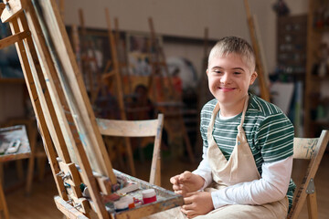 Portrait of young boy with Down syndrome smiling at camera sitting by easel in art class copy space