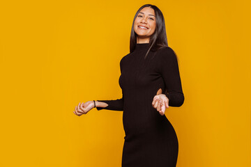 Young latin american smiling elegant woman, wears black dress at studio isolated over yellow...