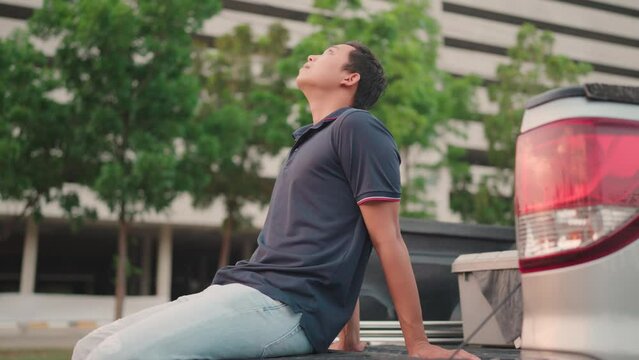 A bored Asian man sits in the back of a pickup truck and looks sideways. Thinking about something or working at home A man who is tired of working far is also boring and lazy.