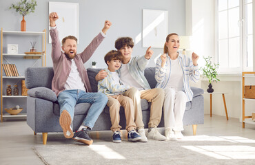 Portrait of young excited happy family of four with children boys watching tv sitting on sofa at home. Parents with two sons supporting football sport team together in living room. Football fans.