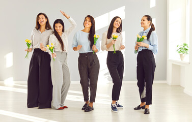 Obraz premium Team of young women with flowers having fun in business workplace on International Women's Day. Group of happy beautiful ladies receive spring tulips at work in office. Holiday, celebration concept