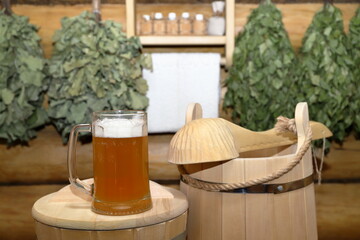 A glass mug of light beer stands on a wooden bucket in the sauna interior on the background of a log wall with dry brooms and shelf with set of aroma oil bottles and white towel. 