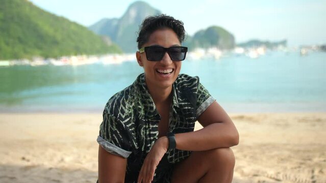 Young girl sits on the soft sands of a sunlit beach, wearing a relaxed patterned shirt, with the gentle waves of a serene sea in the background. The mood is carefree and joyful. Slow motion.